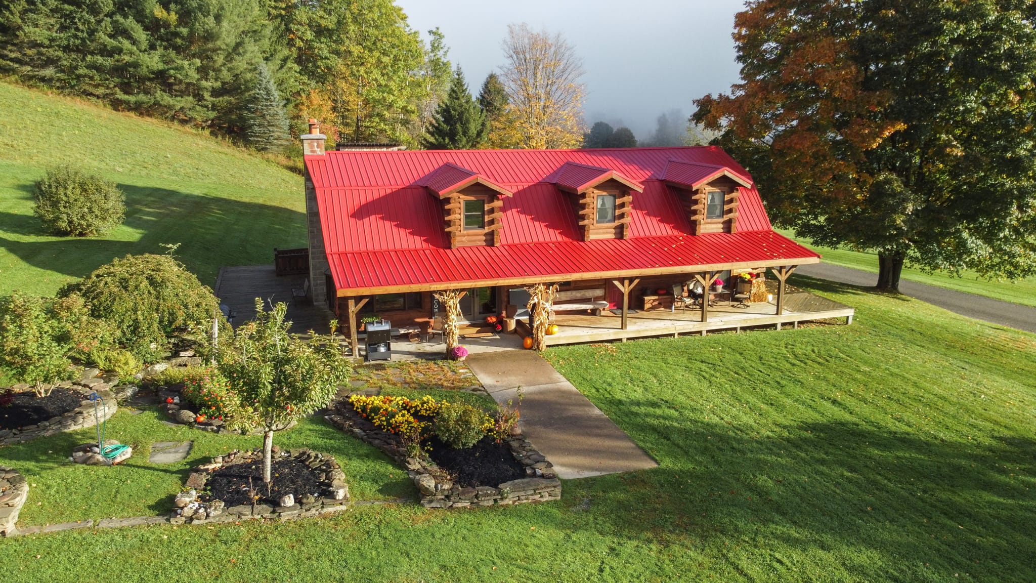 DJI_0585 Clara Creek Company, Rustic cabin with red roof and covered porch, surrounded by a lush green lawn and autumn trees.