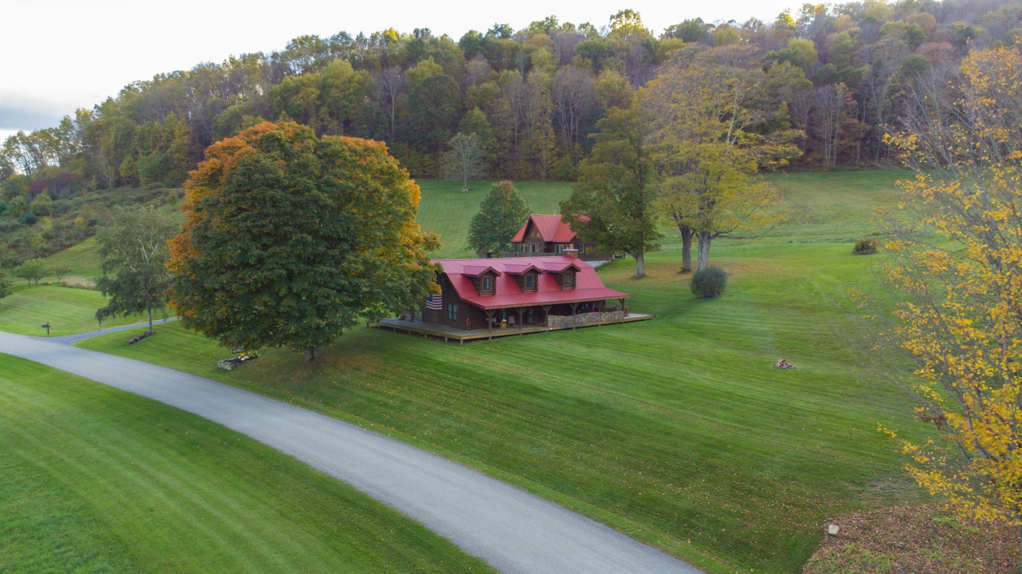 DJI_0492 2 Clara Creek Company, Scenic view of a red-roofed log cabin nestled among autumn trees and a green lawn.