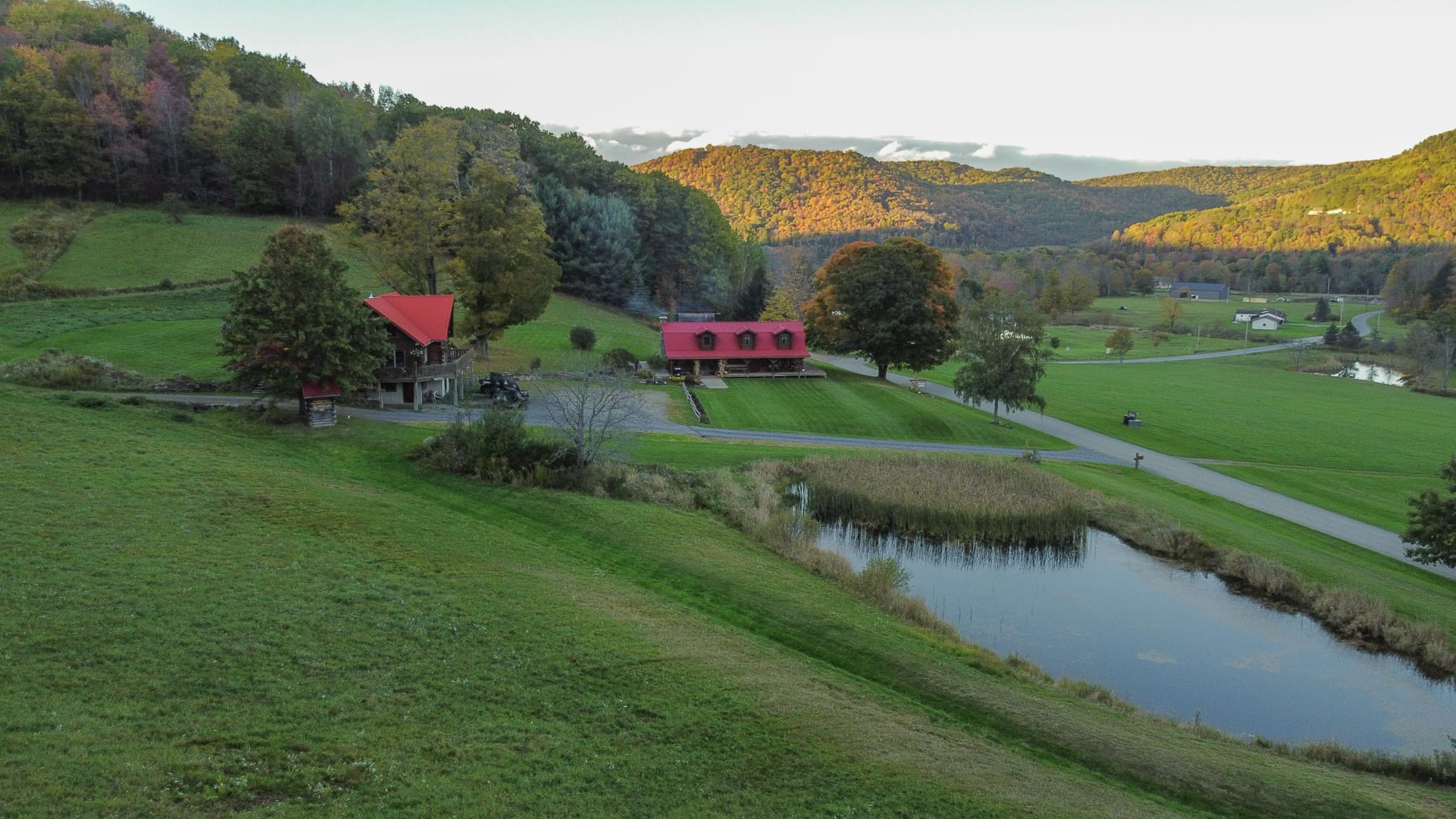 DJI_0562 Clara Creek Company, Scenic view of two red-roofed houses nestled in a green valley with a pond and autumn foliage.